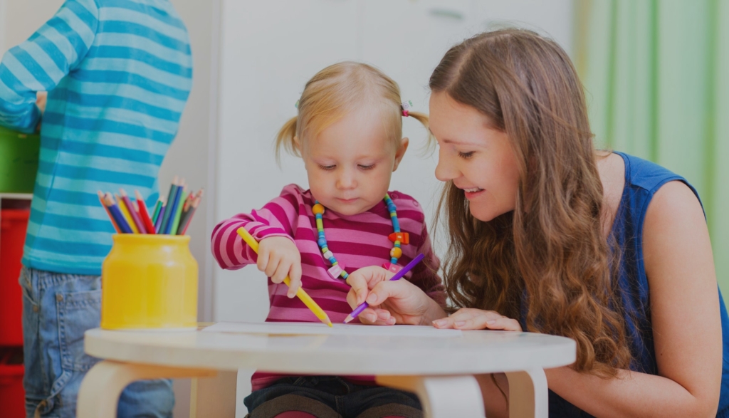 A little girl coloring with the help of a smiling teacher.