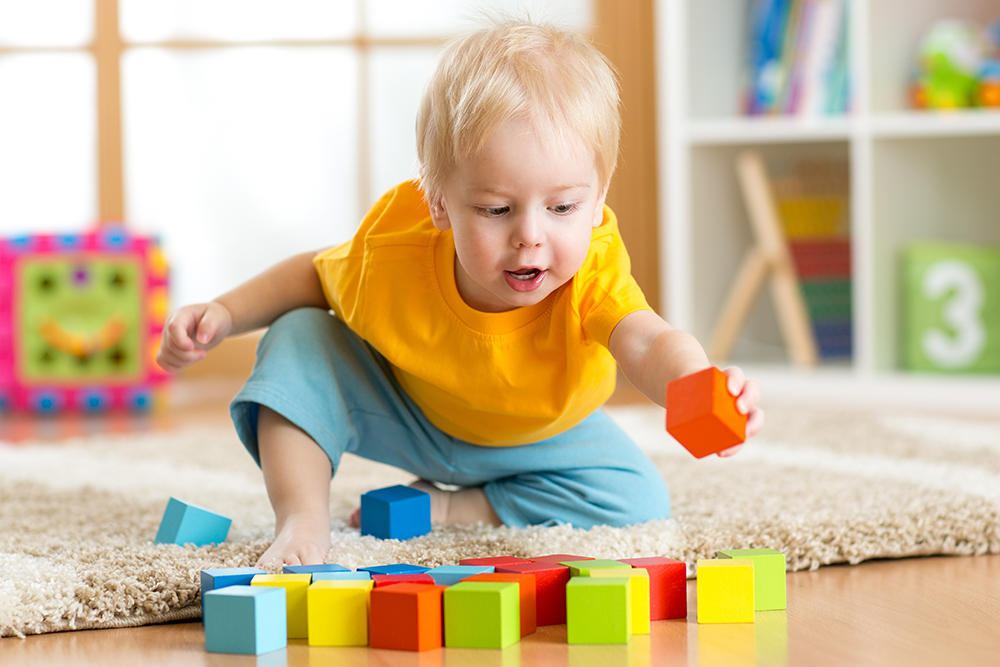 A blonde toddler reaches for colorful blocks while playing on the floor.