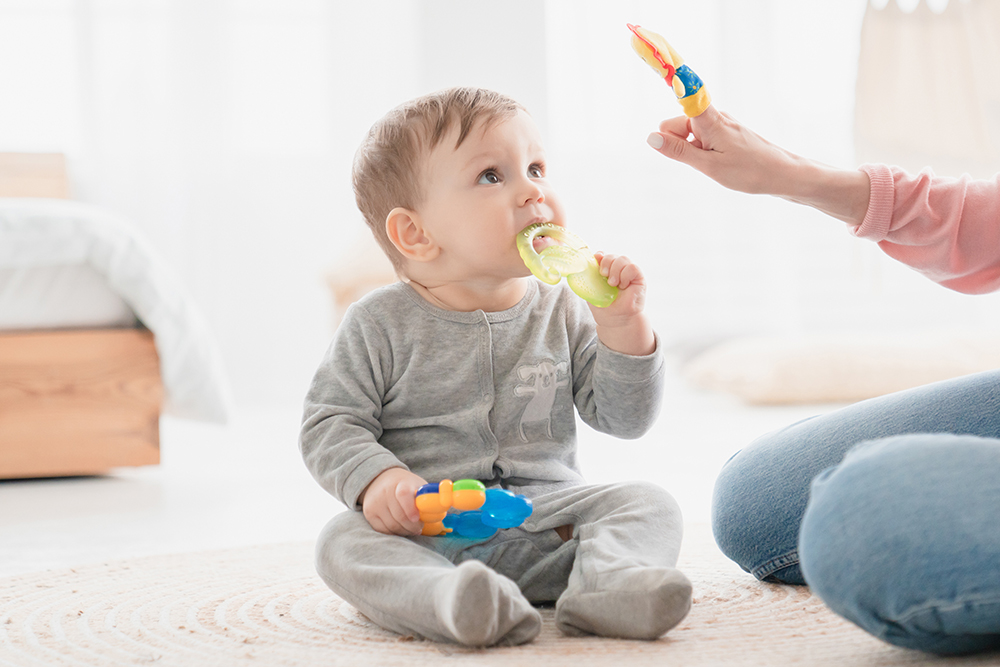 A baby in grey pajamas sits on the floor and chews on a green teething toy while looking up.