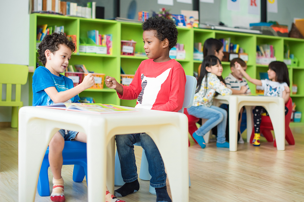 Children sitting at small tables in a classroom, talking and interacting with each other.