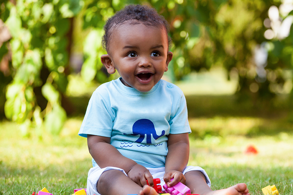 A happy baby sits outdoors on the grass, playing with colorful toys in the sunshine.