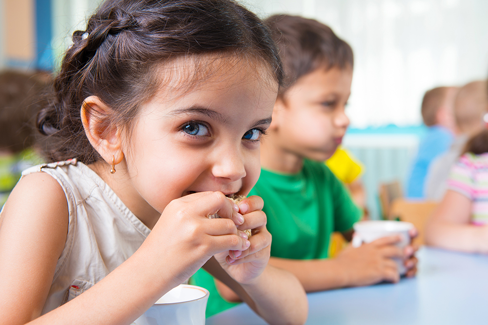 A young girl eating food while sitting at a table with other children.