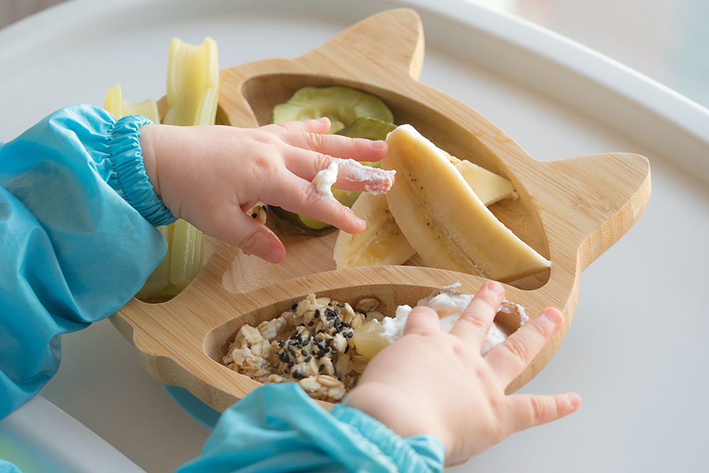 A baby’s hands explore food on a wooden cat-shaped plate during a meal.
