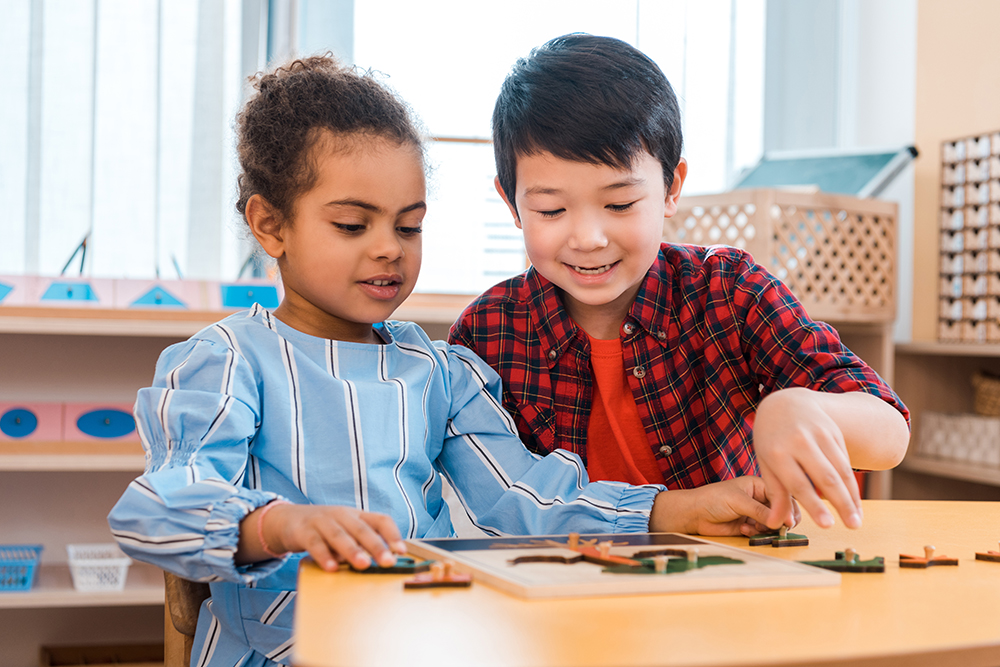 A boy and a girl work together on a wooden puzzle at a classroom table.