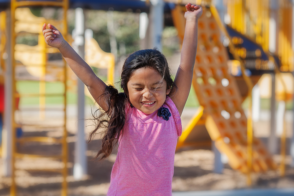 A happy girl lifts both arms in excitement at an outdoor playground.