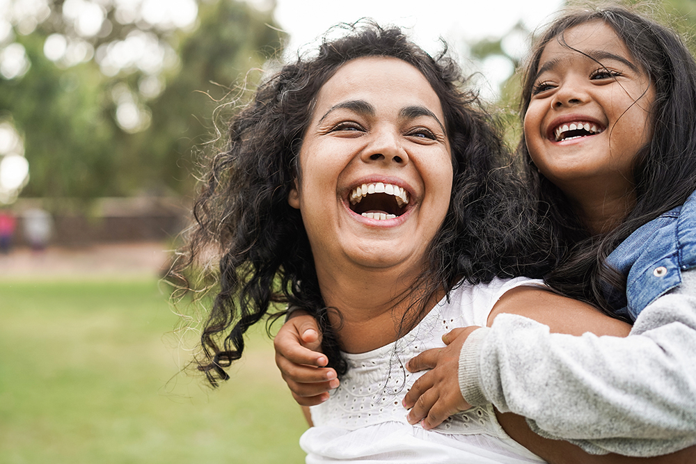 A teacher laughing while carrying her daughter outdoors.