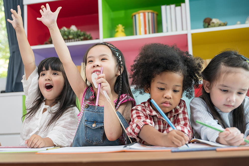 Four children sitting together at a table, two raising their hands and two writing.