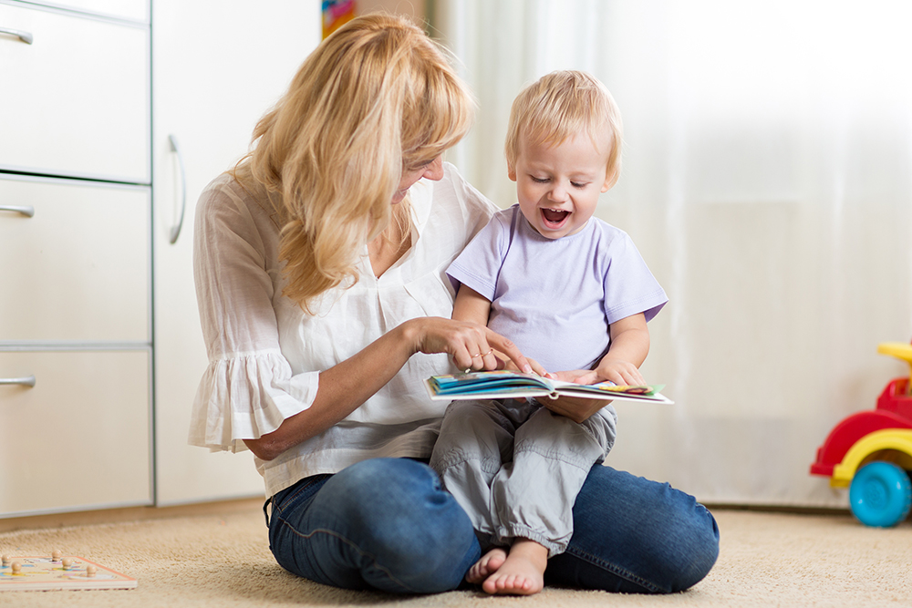 A teacher sits on the floor reading a book with a delighted toddler on her lap.