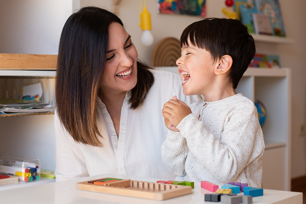A smiling teacher and child laugh together while playing with small colorful educational blocks.