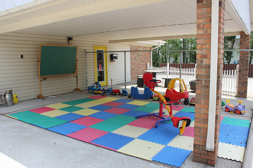 A covered outdoor space with colorful floor mats, ride-on toys, and a chalkboard.