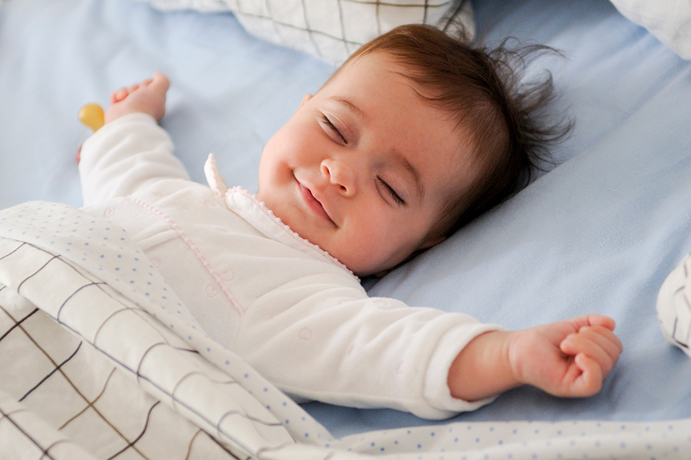 A baby sleeps comfortably in bed with arms stretched out and a slight smile.