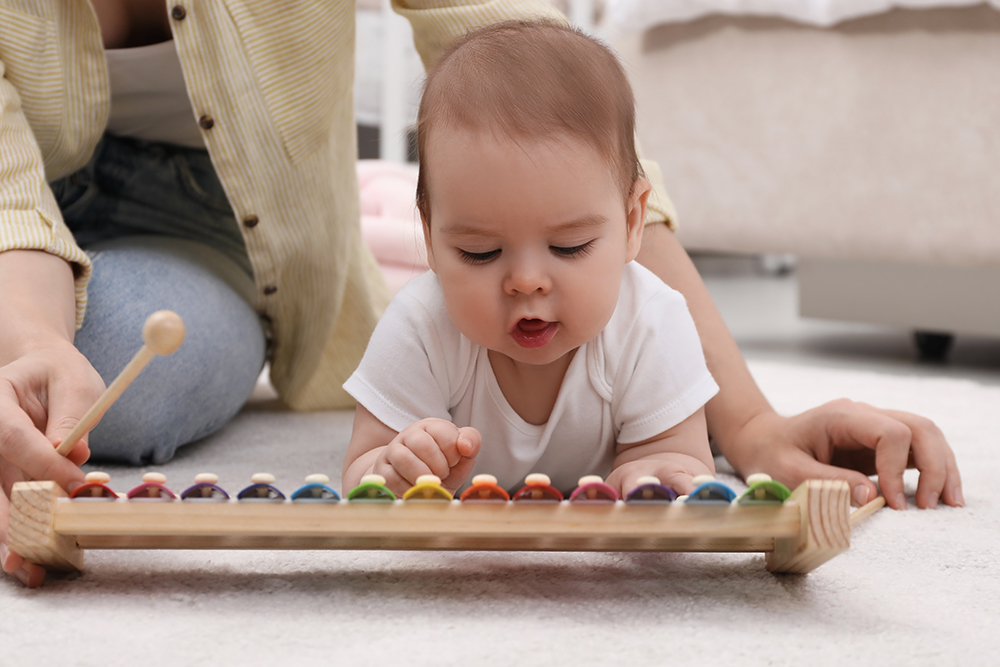 A baby lies on their belly, exploring a colorful wooden xylophone while an adult helps.